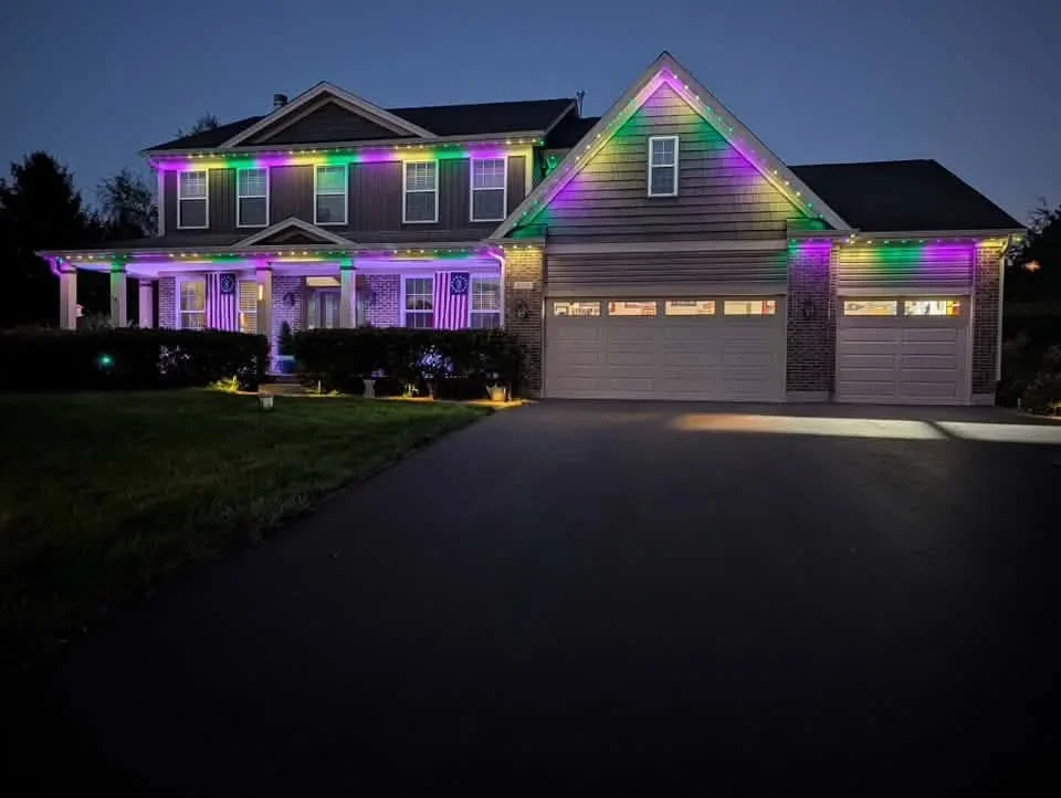 Two-story brick home with multi-color LED holiday lights on rooflines, gables, and garage doors at dusk