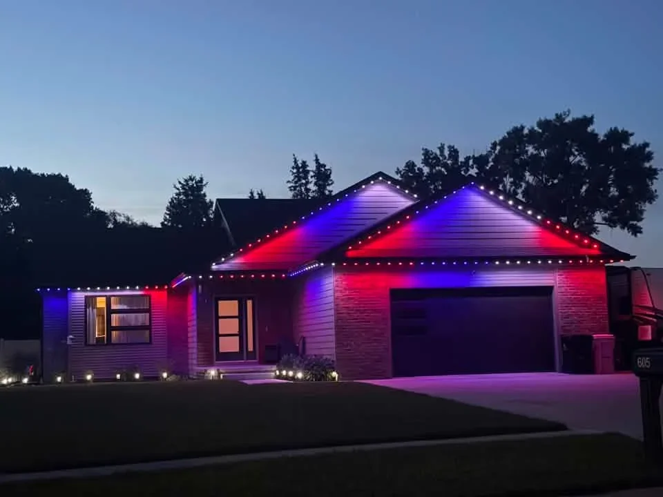 Modern home with RGB LED roof and soffit lighting in red and blue, ground-level landscape lights illuminated at dusk