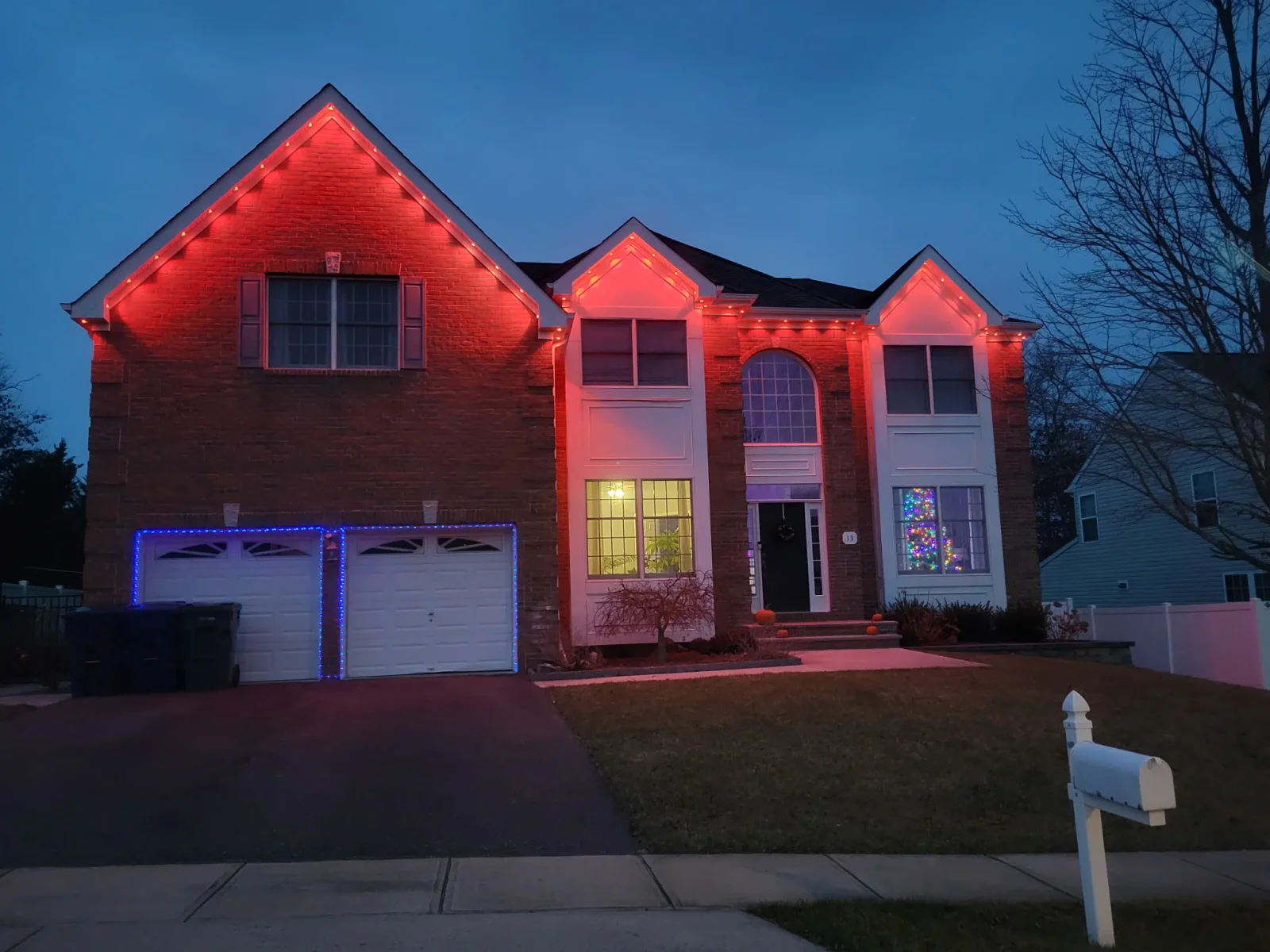 Two-story brick home with red roofline lighting, blue garage door accent lights, and warm interior window lighting at dusk
