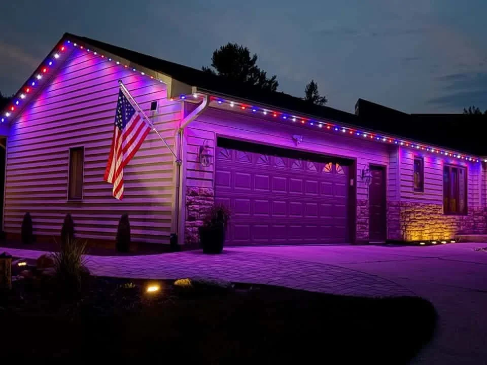Garage exterior with purple RGB LED strip lighting along roofline and stone foundation uplighting at dusk